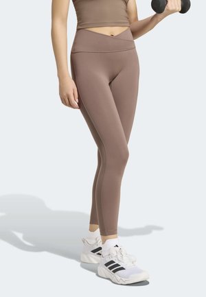 Osoba nosící fialové legíny a hnědý krátký top, zvedá černou činku jednou rukou, stojí v bílých teniskách Adidas.