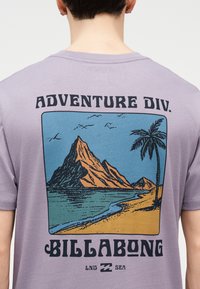 Άτομο που φοράει ένα λιλά μπλουζάκι με γραφικό που απεικονίζει βουνό, φοίνικα, παραλία και πουλιά, με το κείμενο «ADVENTURE DIV. BILLABONG» στην πλάτη.