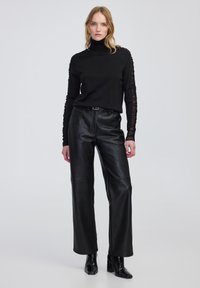 Vrouw in een zwarte coltrui met kantmouwen, zwarte leren broek en zwarte enkellaarsjes met hak, staand tegen een effen achtergrond.