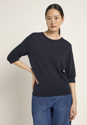 Navy pullover aus Rippstrick mit rundem Ausschnitt und Dreiviertelärmeln, kombiniert mit hellblauen Jeans. Weicher Stoff und lässige Passform.
