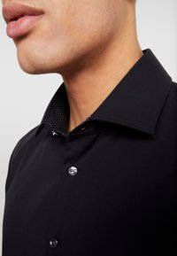 Närbild av en mans nacke och haka som bär en svart skjorta med knappar och en inre krage med svart och vit prickig mönstring.