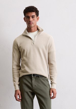 Beige rits-pullover met textuurstof, geribde manchetten en kraag, gecombineerd met olijfgroene broek. Model staat tegen een neutrale achtergrond.