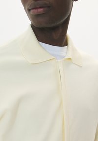 Cămașă polo de culoare galben pal, cu fermoar frontal, guler scurt și un material texturat moale, purtată peste un tricou alb.