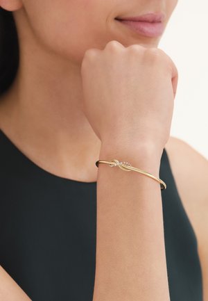 Primo piano del polso di una donna che indossa un sottile braccialetto d'oro con un nodo e piccole pietre trasparenti, su una maglia nera senza maniche.