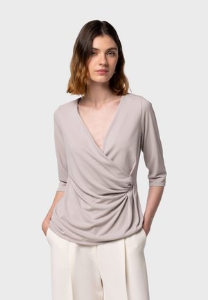 Light taupe wrap top with a deep V-neck, three-quarter sleeves, and draped texture, paired with cream trousers. Smooth, stretchy material.