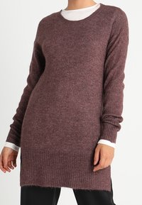 Maglione a maglia color bordeaux con maniche lunghe e polsini a costine. Tessuto morbido, scollatura a girocollo, indossato sopra una maglietta bianca.