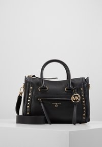 Borsa Michael Kors in pelle nera con texture, borchie dorate, tasca con zip frontale, doppi manici e tracolla staccabile su superficie bianca.