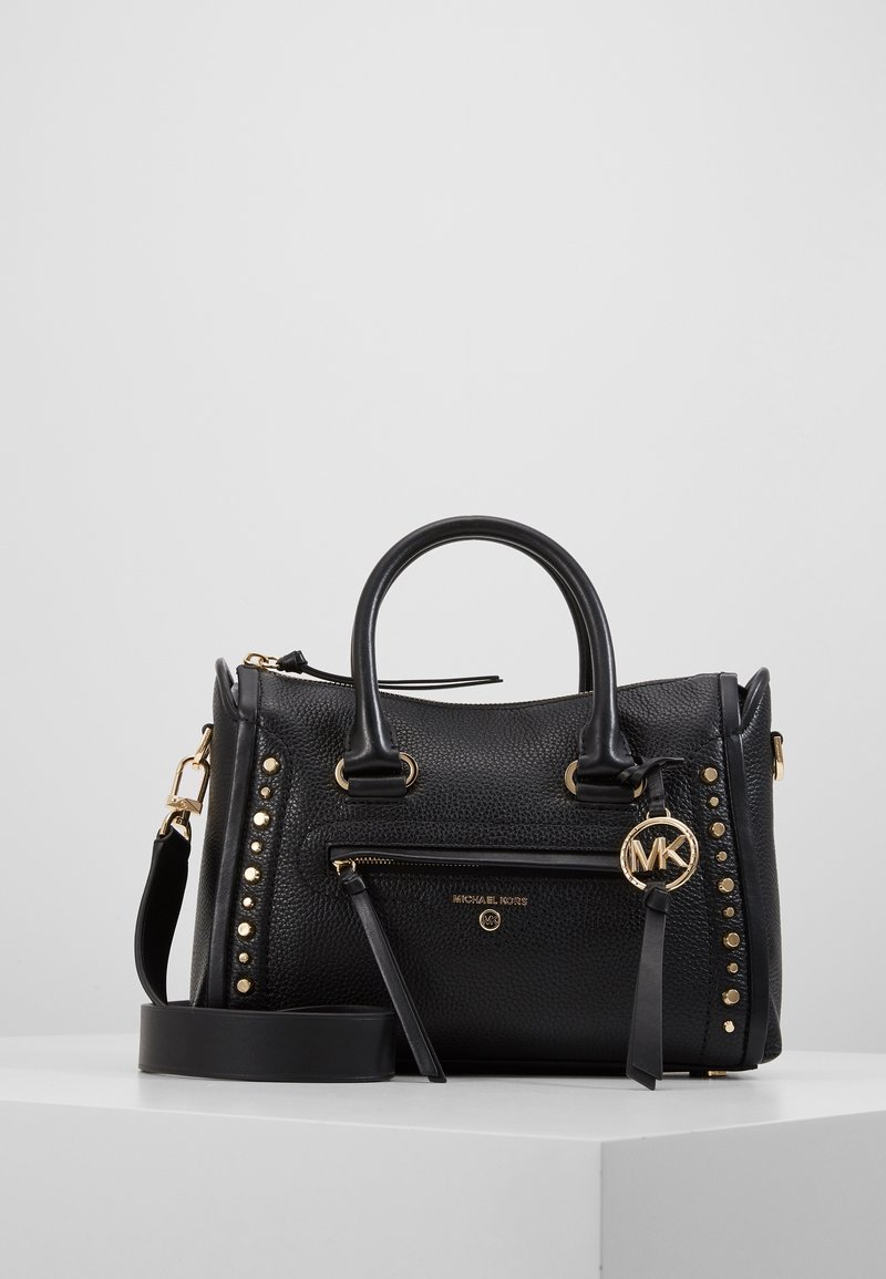 Borsa Michael Kors in pelle nera con texture, borchie dorate, tasca con zip frontale, doppi manici e tracolla staccabile su superficie bianca.