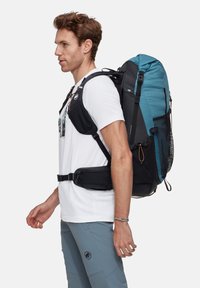 Ryggsäck med blå topp och svart botten, utrustad med nätfickor och justerbara remmar, buren av en man i vit skjorta och grå byxor.