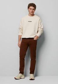 Cremefarbener Sweatshirt mit schwarzem "JIL SANDER"-Logo, braune Slim-Fit-Hose und beigefarbene Sneaker mit roten Akzenten. Strukturiert und lässig im Design.