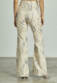Beige high-waisted broek met blauwe bloemen- en vogelborduurwerk, gedragen met lichtblauwe hoge hakken op een betonnen vloer.