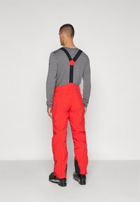 Mono de esquí rojo con tirantes negros ajustables, piernas ajustadas y costuras reforzadas. Combinado con una camiseta gris de manga larga y botas negras.