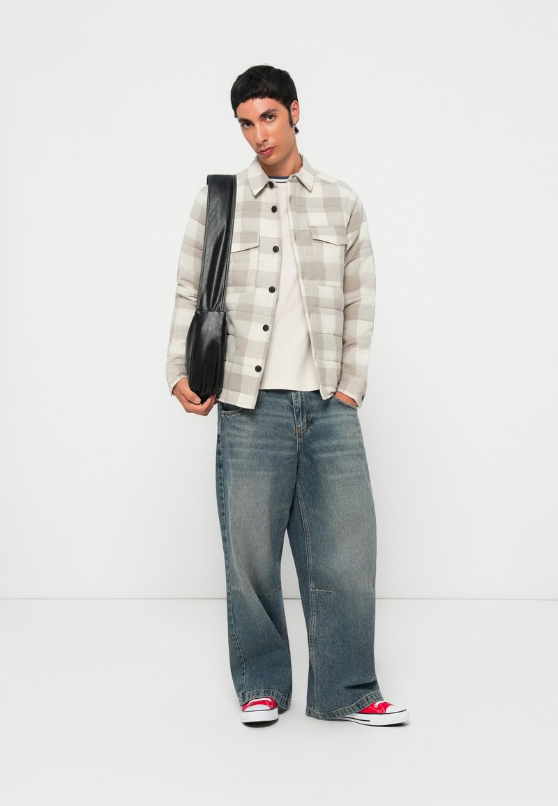 Junge Person steht mit einem beige karierten Jackett, einem weißen Hemd, weiten blauen Jeans, roten Turnschuhen und trägt eine schwarze Umhängetasche.