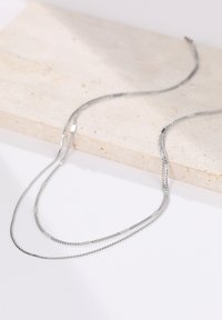 Silbernes Doppelstrang-Halsband mit einer feinen Kette und rechteckigen Akzenten, auf einer strukturierten Steinoberfläche präsentiert.