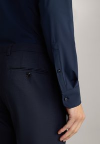 Marineblauwe shirt met lange mouwen, voorzien van knoopmanchetten. Gecombineerd met donkerblauwe broeken met een zak met een knoopdetail.