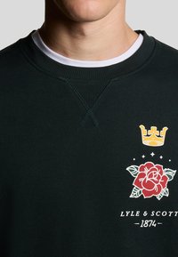 Dunkelgrüner Sweatshirt mit geripptem Kragen, das ein weiß gesticktes Kronensymbol und ein rotes Rosengrafik mit grünen Akzenten sowie Logotext zeigt.