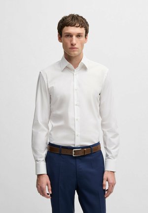 Chemise blanche à manches longues avec un col classique, fermeture à boutons et manches à poignets. Portée avec un pantalon bleu marine et une ceinture en cuir marron.