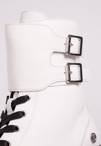 Botín de piel blanca con cordones negros, que presenta dos hebillas metálicas, textura suave y diseño minimalista. Logo de acento en el lateral.