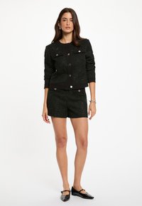 Svart texturerad jacka med silverknappar, matchande skräddarsydda shorts och svarta platta skor. Har en rund halsringning och två framfickor.