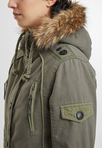 Olijfgroene parka met een nepbont capuchon, trekkoorddetails, rits- en klepzakken, en schouderaccenten. Getextureerde stof.