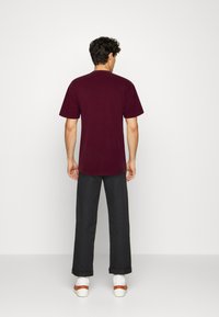 T-shirt a maniche corte, color bordeaux, con scollo rotondo. Indossato con pantaloni larghi scuri e scarpe bianche con cinturini marroni. Design semplice, vestibilità media.