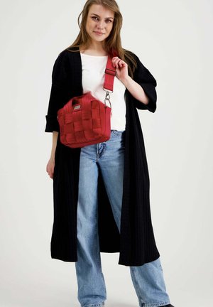 Nainen, jolla on päällään pitkä musta neuletakki, valkoinen paita, siniset farkut, pitää punaisen punotun crossbody-laukun säädettävällä hihnalla vaalealla taustalla.