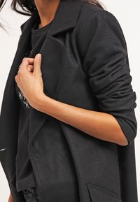 Abrigo de mezcla de lana negra con cuello de solapa, diseño ajustado y mangas arremangadas. La persona que lo lleva lo sostiene abierto, revelando un interior negro.