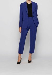 Costume bleu ajusté avec un blazer à un bouton et un pantalon slim. Top noir en dessous. Complété par des talons à bride noirs.