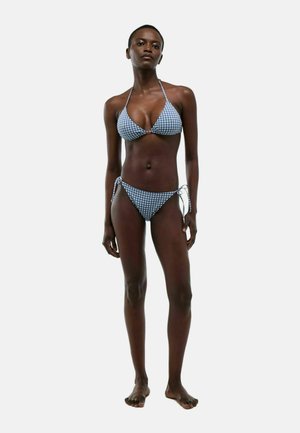 Femme debout portant un bikini vichy bleu et blanc avec des liens sur le haut et les côtés, faisant face à l’avant sur un fond uni.