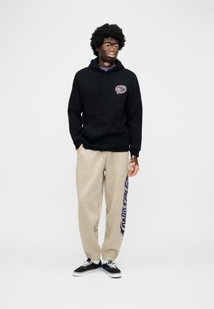 Jonge man met vitiligo, gekleed in een zwarte hoodie met grafisch embleem en beige broek met donkere, patroonrijke strepen, staand tegen een witte achtergrond.