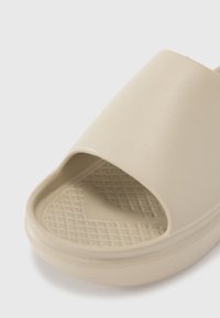 Sandale glissante beige avec une finition lisse et mate, design ouvert à l'avant, semelle intérieure à motif en grille et semelle arrondie. Matériau durable et flexible.