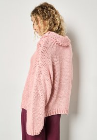 Różowy sweter z dzianiny z kapturem, o luźnym kroju i teksturowanym ściągaczu. Długie rękawy, opadające ramiona oraz krótki krój.