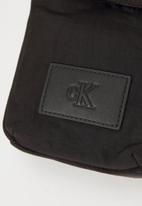 Borsa a tracolla nera in nylon con una patch in pelle nera goffrata con il logo "cK". Texture liscia; design minimalista.