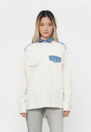 Vrouw met lang haar draagt een oversized witte blouse met knopen en een denim kraag, schouderaccenten en contrasterende zakken, gecombineerd met lichtgrijze broek.