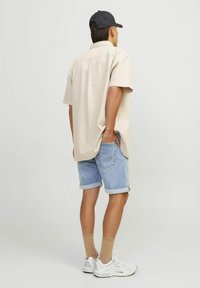 Man wearing beige short-sleeve shirt, light blue denim shorts, beige socks, white sneakers, and a black cap standing with one hand in pocket.