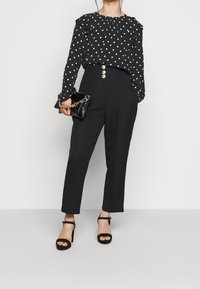 Blusa nera a pois con spalline arricciate, pantaloni neri a vita alta con bottoni dorati e decolletè neri con cinturino alla caviglia. Clutch nera.