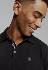 Zwarte poloshirt met een gestructureerde afwerking, voorzien van een klein hexagonaal logo op de patch en lichtblauwe details in de kraag.