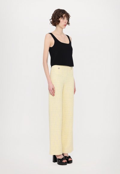 Sort cropped tank top, gule strukturerede bukser med bred ben og dobbelte frontknapper, samt sorte platform sko med sløjfe accenter.