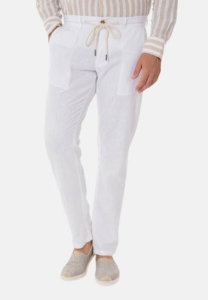 Uomo che indossa pantaloni di lino bianchi con coulisse, camicia beige e bianca a righe e scarpe slip-on grigio chiaro, in piedi con le mani nelle tasche.