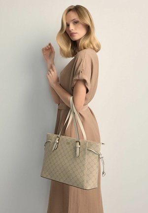 Blondynka w beżowej sukience trzyma dużą beżową torebkę z wzorem i podwójnymi paskami na ramiona, stoi na tle gładkiej, jasnej ściany.