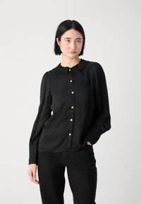 Blouse noire à manches longues avec un motif léopard subtil, un col en dentelle et des boutons carrés. Tissu lisse avec une coupe décontractée, associée à un pantalon sombre.