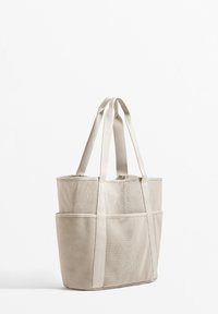 Beige hálós bevásárlótáska dupla fogantyúkkal és több külső zsebbel, téglalap alakú és texturált felülettel.