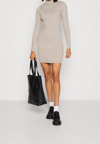 Abito lavorato a maglia beige con una silhouette aderente, maniche lunghe, texture a coste, abbinato a una borsa tote nera e scarpe slip-on nere.