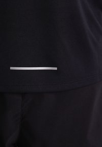 T-shirt de sport noir avec une texture lisse, des manches courtes et une fine bande réfléchissante horizontale argentée près de l'ourlet.