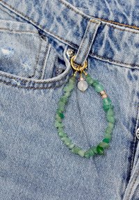 Pärlbeväpnad nyckelring med gröna stenar i olika former, gulddetaljer och en transparent pärla, fäst vid en jeansficka.