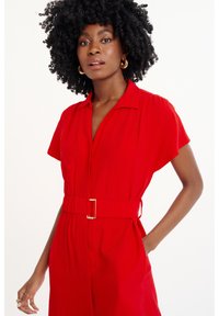 Rotgestreifter Jumpsuit mit kurzen Ärmeln, kragenlosem Ausschnitt und zwei seitlichen Taschen. Der Stoff wirkt glatt und leicht, mit einer taillierten Passform.