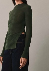 Haut à manches longues côtelé vert foncé avec un col haut, des boutons sur le côté et un ourlet légèrement fendu, associé à un jean foncé.