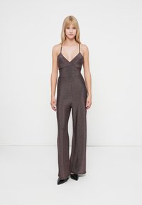 Salopetă strălucitoare, de culoare roz închis, cu bretele subțiri, corset ajustat, picioare evazate și o textură subtilă. Pantofi negri cu vârf ascuțit completează ținuta.