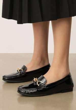 Jambes portant des mocassins noirs en cuir verni brillant avec un détail de chaîne dorée, assortis à une jupe plissée noire.