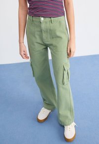 Groene wijde cargobroek van katoen, met zijzakken en een hoge taille, gecombineerd met witte sneakers op een blauwe achtergrond.